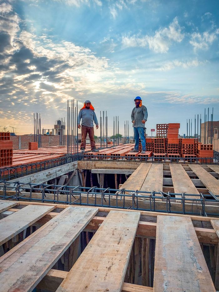 Two construction workers stand on a building site under a cloud-filled sky. Sunlight illuminates the scene.